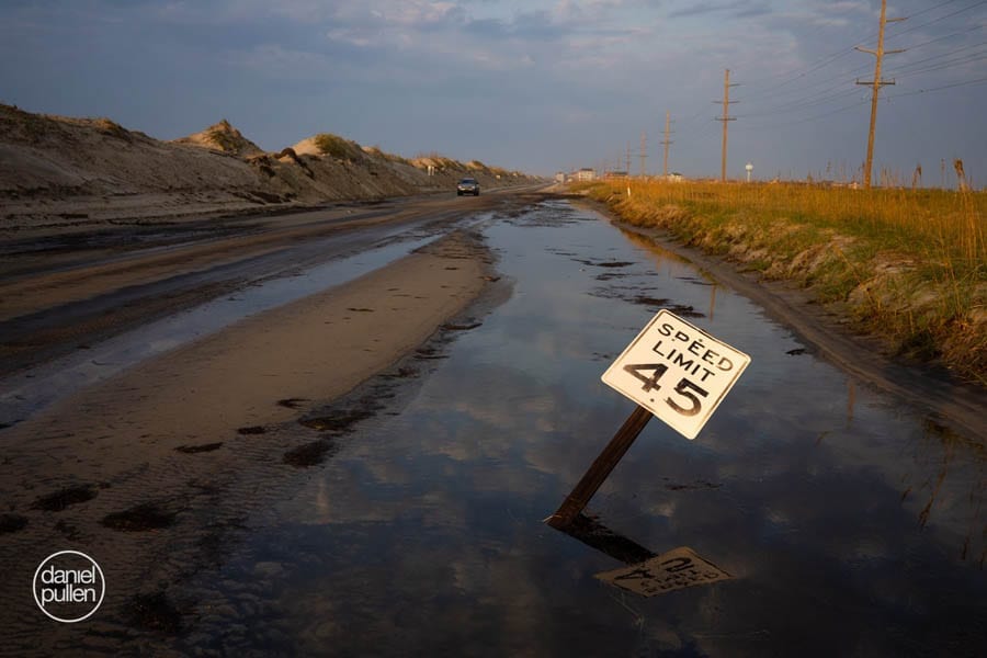 Image of N.C. 12 roads covered in sand and water after a hurricane. A 45mph speed limit sign leans toward the ground.