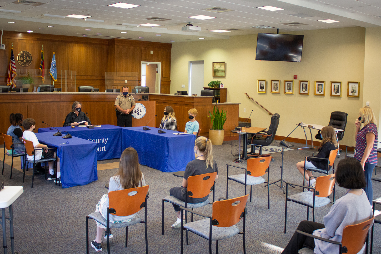 Image of teen court trial. Four students and a judge sit at a table while other students watch from chairs.